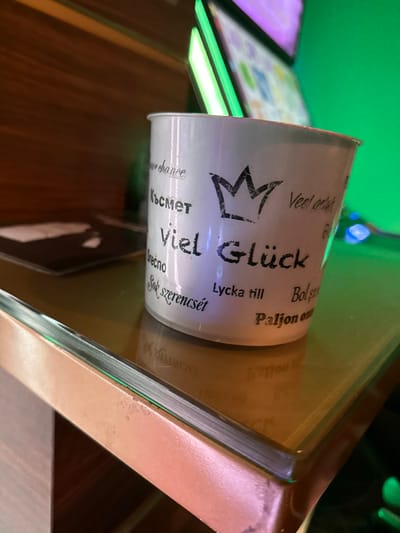 The image displays a close-up, angled view of a white cylindrical mug, possibly made of ceramic or plastic, positioned on a reflective, brownish-tinted glass surface. The mug's visible side features the prominent black text "Viel Glück" (German for "Good Luck") surmounted by a stylized, distressed black crown graphic. Around these central elements, various phrases are printed in different languages, including "Късмет" (Bulgarian), "Sok szerencsét" (Hungarian), "Lycka till" (Swedish), and "Paljon onnea" (Finnish), all conveying wishes of luck or happiness.

The supporting glass surface has a discernible copper-toned or reddish-brown metallic trim along its visible edge. Reflections of the mug's text are faintly visible on this surface. The background is out of focus, revealing a dark wooden surface, likely part of furniture, and a bright green light source or illuminated screen, from which indistinct rectangular reflections are cast. No individuals are present in the scene, and no specific actions or interactions are occurring. The visual evidence in the image does not provide identifiable markers to confirm its location in Lohfelden, Germany.