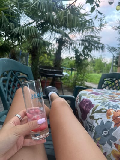 The scene depicts an individual in Isperihovo, Bulgaria, seated outdoors in a green plastic chair, seen from a first-person perspective, with legs extended. The subject's right hand, with light purple nail polish and a silver ring on the ring finger, holds a clear drinking glass containing a pink liquid and what appears to be ice or fruit pieces; blue text, possibly "ZAK," is visible on the glass. The subject's left foot, wearing a black slide sandal with a white sock, is also visible. To the right, a table covered with a floral tablecloth featuring patterns of purple, blue, orange, and green flowers on a white background is present. In the mid-ground, a black barbecue grill is visible to the left. The background consists of out-of-focus green foliage, including prominent palm trees, under an overcast sky.