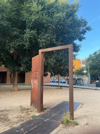 The image depicts an outdoor scene in Figueres, Spain. The primary focus is an abstract metal sculpture that resembles an open doorway, constructed from rusted metal. One vertical element of the sculpture leans against a large, reddish-brown plaque inscribed with text, some of which is obscured by grime and scratches. The text appears to be in multiple languages. This plaque is positioned on the ground, extending forward from the base of the sculpture.
The ground is covered in sand and dirt, with sparse vegetation and small yellow wildflowers growing around the base of the sculpture and plaque. Behind the sculpture, a large, leafy tree with a thick trunk dominates the left side of the frame, providing shade. In the background, there is a brick building with arched entrances and signs, suggesting it might be a commercial establishment. Further in the background, a street is visible with cars parked and driving, along with large grey trash receptacles. The architecture in the distance includes buildings with orange and yellow facades. The sky is a clear, bright blue with a few wispy clouds. The overall impression is a public space, possibly a park or plaza, with an artistic installation as its centerpiece.