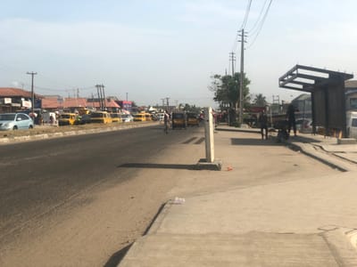 An outdoor, wide-angle street view in Alasia, Nigeria, depicts a daytime urban scene under a clear sky. A two-lane asphalt road, appearing worn, occupies the foreground and mid-ground, receding into the distance. A concrete sidewalk runs along the right side of the road, featuring a metal-framed bus stop shelter without a roof covering. Multiple wooden utility poles with overhead wires are visible on both sides of the street.
On the left side of the road, a dirt median separates the main thoroughfare from a paved shoulder where a light blue sedan is parked. Behind it, a queue of at least seven yellow mini-buses, commonly known as danfo, are either parked or slowly moving. Several individuals are visible on the sidewalk and shoulder adjacent to these vehicles.
On the right side of the road, near the bus stop, four individuals are present: one person stands near the shelter, another is partially obscured behind it, and two are closer to the foreground on the sidewalk, one standing and one in a crouched position. A white van is partially visible behind the bus stop structure. Closer to the foreground on the right, a concrete bollard stands on the median, with some red and white debris on the ground nearby.
Further down the main road, one person walks towards the right side, and a three-wheeled vehicle (tricycle) is in motion. Buildings with reddish-brown roofs and various signs line the left side of the street in the background. Sparse trees and vegetation are scattered throughout the scene. The lighting indicates direct sunlight, casting shadows.