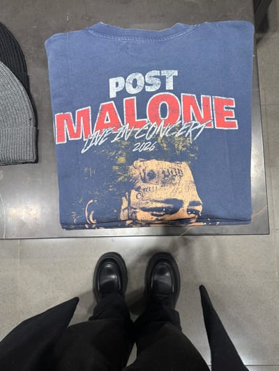 A navy blue t-shirt with "POST MALONE LIVE IN CONCERT 2026" printed in red and white sits on a gray surface. The shirt features an image of Post Malone's face. Below the shirt are two knit hats, one black and one gray. A person wearing black pants and black loafers stands next to the surface, with the tips of their black coat visible. The surface appears to be a table or display in a retail setting.