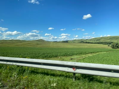 The image depicts a rural landscape under a bright blue sky with scattered white clouds. The foreground features a gray metal guardrail, slightly blurred due to motion, with green grass and some wildflowers growing beside it. Beyond the guardrail, a vast field of vibrant green crops stretches towards rolling hills in the background. The hills are covered in green vegetation, with some trees scattered across the landscape. The scene appears to be a view from a vehicle traveling along a road, indicated by the blur in the foreground and the perspective. The location is identified as Isyangulovo, Russia.