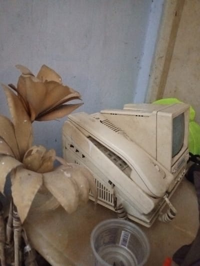 The image depicts an indoor scene in Keo Kouang, Laos, featuring a collection of aged objects on a light brown surface against a light-colored wall. In the left foreground, a large, dusty, faded beige artificial plant with multiple layered petals is prominently displayed. To its right, in the midground, an off-white combination telephone and fax machine is visible, with its handset resting on the main unit, revealing a numeric keypad and ventilation grilles. Immediately behind or stacked upon this device is a compact, off-white CRT monitor or television. A clear plastic cup with a barcode label is situated in the lower right foreground, containing a small amount of dark liquid or debris. The background consists of a light grey, textured wall showing signs of dust and discoloration, with a vertical wooden beam or frame partially visible on the far right. A bright, possibly neon green fabric or object is partially visible behind the electronic devices on the right.