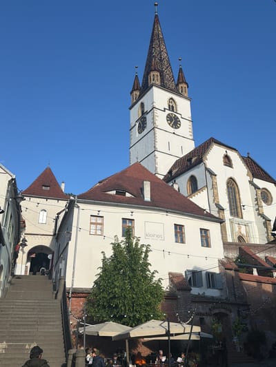 The image depicts a view of Sibiu, Romania, with a prominent church tower featuring a decorative roof and clocks on its white facade. Adjacent to the tower are buildings with red tile roofs and white walls. One building has a sign for "Kleines Hotel." A tree partially obscures the view. The foreground shows stairs leading up to an area with tables and chairs, suggesting an outdoor cafe or restaurant. People are seated at some of the tables. The sky is clear and blue.