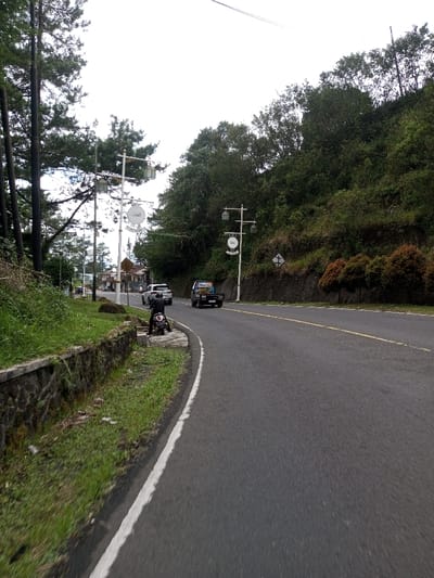 The image depicts a two-lane asphalt road curving gently to the right through a hilly, vegetated area in Phoxay, Laos. The sky is overcast.

On the left side of the road, a low stone retaining wall with a concrete cap separates the asphalt from a strip of green grass and dirt. A person wearing a dark jacket and helmet is seated on a dark-colored motorcycle, parked on this grassy shoulder, facing away from the viewer. Behind the retaining wall and motorcycle, tall evergreen trees line the left edge of the scene. Multiple white, decorative streetlights with lanterns and circular signs are spaced along this side; one prominent sign displays Arabic script, reading "العدل" (Al-Adl, meaning Justice). Buildings are visible in the distance beyond these trees and streetlights.

The road features a solid white line marking its left edge and a solid yellow line indicating the center. Traffic is flowing away from the viewer. A dark-colored pickup truck with an open bed is traveling in the right lane, followed by a white SUV further ahead.

The right side of the road borders a steep, heavily vegetated hillside, densely covered with green trees and shrubs. Another decorative streetlight with a circular sign (less clear, possibly depicting "السلام" - As-Salam, meaning Peace) is positioned on this side. A white diamond-shaped sign with a black border and a diagonal cross is affixed to the hillside. Further down, a row of cultivated reddish-brown shrubs is planted along the base of the hill. Overhead, power lines are faintly visible against the sky.