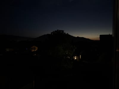 The image depicts a dark, nighttime landscape with subtle hints of twilight on the horizon. Silhouetted against the dusky sky are rolling hills and mountains. In the foreground, the shapes of trees and buildings are barely discernible in the low light. A few scattered lights from houses are visible, suggesting a rural or suburban setting. The overall impression is one of stillness and quietude. The location is identified as Vyzhnytsia, Ukraine. There are no people or animals present in the image, and no actions or interactions are depicted.