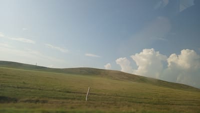 The image depicts a rolling green hillside under a pale blue sky. A single, thin utility pole stands prominently on the crest of the hill in the distance, with a faint path visible leading up to it. The foreground shows a field of dry grass, with a wooden fence post visible on the left side. Several large, fluffy white clouds are scattered across the sky, particularly on the right side of the frame. The reflection of a vehicle's interior, including a dashboard and possibly a passenger's headrest, is visible in the upper right portion of the image, suggesting the photograph was taken from inside a moving vehicle. The landscape appears to be rural, possibly in the vicinity of Krasnoschekovo, Russia, given the context.