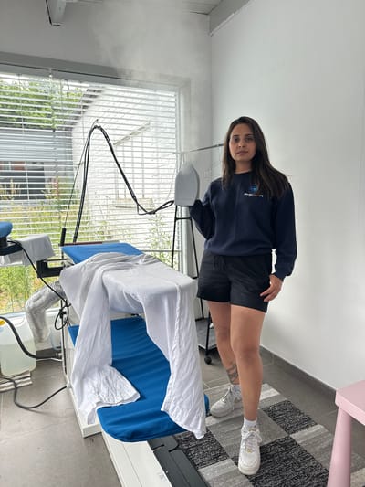A woman stands in a room in Maldegem, Belgium, holding a steam iron over an ironing board. The woman is wearing a dark blue sweatshirt with a logo, black shorts, and white sneakers. A white sheet is draped over the ironing board, and a large plastic container of water is on the floor next to it. The room has a large window with blinds, and there is a grey and black patterned rug on the floor. Steam is visible in the air near the window.