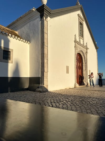Exterior shot of a church in Vila Real de Santo António, Portugal, taken during daylight hours. The church's facade is predominantly white with a gray roof and stone accents around the windows and a wooden door. Two individuals stand near the door, one appearing to take a picture of the other. A cobblestone pavement extends alongside the building, with a curb in the foreground. The sky is clear and blue.