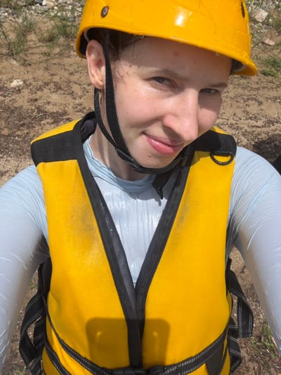 This is a close-up, top-down shot of a young person wearing a yellow safety helmet and a bright yellow life vest. The person is looking upwards, with their face partially obscured by the helmet and the life vest straps. Their skin appears slightly flushed and possibly sweaty, suggesting they might be engaged in an outdoor activity. Their lips are slightly parted, giving a hint of a smile.
The setting appears to be outdoors, on a dirt and gravelly ground with some sparse green vegetation visible. The lighting suggests it is daytime, likely sunny, as there are distinct shadows cast on the life vest. The overall impression is of someone preparing for or participating in an adventure sport, possibly related to water activities given the life vest. The person's gaze and slight smile convey a sense of anticipation or enjoyment. There is no visible text in the image.