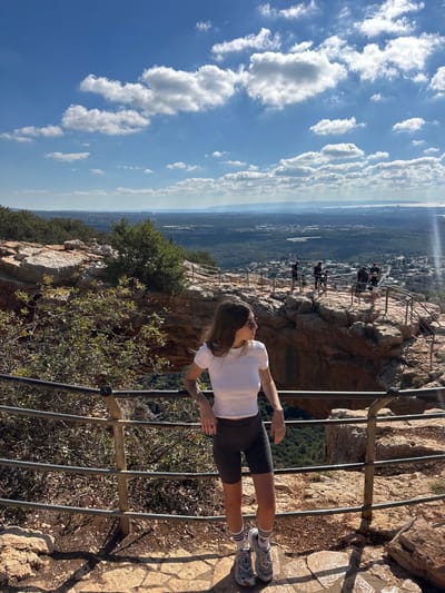 The image captures a panoramic view from a cliffside observation point at Keshet Cave in the Maale Yosef Regional Council, Israel, under a bright blue sky with scattered white cumulus clouds. In the immediate foreground, a female subject stands on a light-colored, irregularly paved stone path. She wears a white short-sleeved crop top, dark gray cycling shorts, white athletic socks with black stripes, and gray sneakers, leaning with both hands on a multi-barred dark metal safety railing. Her brown hair is down, she wears sunglasses, and tattoos are visible on her left arm. Her head is turned to the right, observing the distant landscape.
Behind her and slightly to the right, the distinctive natural rock arch formation of Keshet Cave is visible, with a dark, shaded opening beneath it. Further to the right, on a lower rocky outcrop also equipped with dark metal safety railings, a group of approximately five to six individuals can be seen standing and looking out over the expansive view. The midground and background consist of a vast valley covered with dense green vegetation, scattered buildings, and urban areas. On the distant horizon to the right, a body of water, identifiable as the Mediterranean Sea, is visible alongside what appears to be coastal cities and industrial structures, all under a slight atmospheric haze. Rocks and sparse green bushes are present on the upper left and mid-left portions of the cliff face.