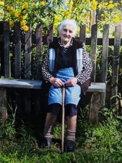 An elderly woman is seated outdoors on a rustic wooden bench in Wojnowice, Poland. She has white hair, fair skin, and facial features consistent with advanced age, looking directly at the camera with a slight smile. She is wearing a black and white patterned long-sleeved blouse, a dark sleeveless vest, a blue patterned skirt, dark leggings or trousers, light brown socks, and dark flat shoes. Her hands are clasped on top of a light brown wooden walking stick positioned vertically between her legs. Behind the woman and the bench is a simple wooden picket fence. Beyond the fence, a dense cluster of bright yellow flowers is visible, thriving under natural sunlight. Green grass grows in the foreground and around the bench, indicating an outdoor, garden-like setting.