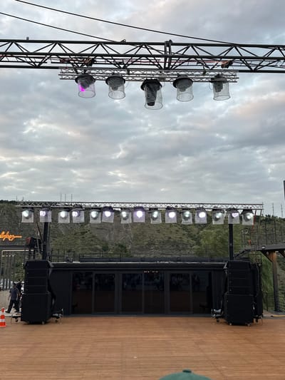 An outdoor stage setup is visible with a wooden floor in the foreground and a cloudy sky in the background. The stage features a black structure with large glass doors, flanked by stacks of black speakers. Overhead, two rows of lighting rigs are suspended from metal trusses. The upper rig holds several large, cylindrical lights covered in clear plastic, with some emitting a pink glow. The lower rig supports multiple smaller stage lights, some of which are illuminated with various colors. To the left of the stage, a neon sign with cursive text is mounted on a metal structure. Several people are present, including two individuals walking on the left side of the image and a group of spectators visible inside the glass-enclosed area of the stage structure. The scene appears to be set in an outdoor amphitheater or event space, with a hillside covered in greenery and trees visible behind the stage. Utility poles and wires are also present in the distance. The overall impression is of a pre-event or setup phase for an outdoor performance.