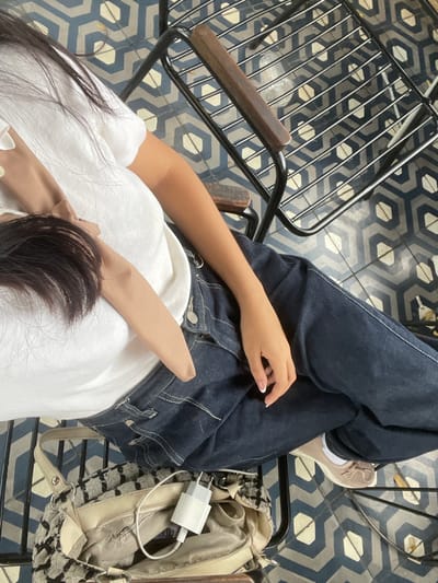 A subject is depicted seated in Limbangan Sari, Indonesia, viewed from an elevated perspective. The individual wears a white short-sleeved top, dark blue denim jeans, and light beige flat shoes. Dark hair is visible across the upper torso. The right hand, featuring a French manicure, rests on the right thigh. The subject occupies a black metal chair with wooden armrests. Adjacent to the subject's left, a beige handbag with a black and white textured pattern is partially open, containing a white USB power adapter and cable. The flooring consists of geometric tiles patterned with repeating blue and light-colored hexagonal shapes.