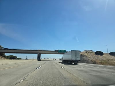 This media file captures a view from inside a vehicle traveling on a highway. The scene depicts a bright, sunny day with a clear blue sky. A wide, multi-lane highway stretches ahead, with concrete barriers lining the sides and intermittent reflective markers on the road.
A prominent element is an overpass with vehicles driving on it, supported by large concrete pillars. To the right of the highway, a dry, grassy hill rises, dotted with sparse bushes. A white semi-truck is in the right lane, moving in the same direction as the viewer's vehicle.
Overhead, a green highway sign is visible. It indicates "Redding" with exit number "5" and "Los Angeles" with exit number "86". Below this, it reads "Jct 99 Mls". This text provides crucial location cues, suggesting the vehicle is on a highway in California, likely heading towards or passing through areas that connect to these cities.
Further down the highway, several other vehicles, including trucks and cars, are visible, indicating moderate highway traffic. The overall impression is a typical day of highway travel in a warm, dry climate, likely California given the signage. The time of day appears to be midday due to the strong sunlight and shadows. No people are clearly visible, reinforcing the perspective from within a vehicle.