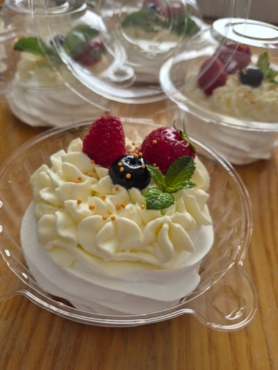 The media file features several individual desserts, likely pavlovas or cream puffs, presented in clear plastic containers. The main subject is a single dessert in the foreground, showcasing a swirl of white cream topped with fresh berries, including a bright red strawberry, a raspberry, and a dark blueberry. A sprig of fresh mint garnishes the dessert, and small, golden sprinkles are scattered across the cream.
The desserts are arranged on a wooden surface, suggesting a rustic or natural setting. The containers are designed with rounded edges and a decorative rim, possibly resembling animal ears, adding a playful touch. In the background, other similar desserts are visible in their containers, some slightly out of focus, indicating they are part of a larger offering or a display.
The lighting is bright and natural, casting soft shadows and highlighting the textures of the cream and berries. The overall impression is one of freshness, sweetness, and delicate presentation. The image evokes a sense of indulgence and simple pleasure, suggesting a treat enjoyed perhaps at a cafe, bakery, or a special occasion. There is no visible text in the media file. The location context of Šilutė, Lithuania, hints at a European setting for this culinary presentation.