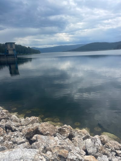 The image shows a body of water next to a rocky shoreline, with a building structure in the distance and a mountain range in the background under a cloudy sky. The location is likely near Sv. Konstantin, Bulgaria. The water appears dark with reflections of the sky and distant mountains. Rocks are clustered along the edge of the water. The building in the distance is a concrete structure.