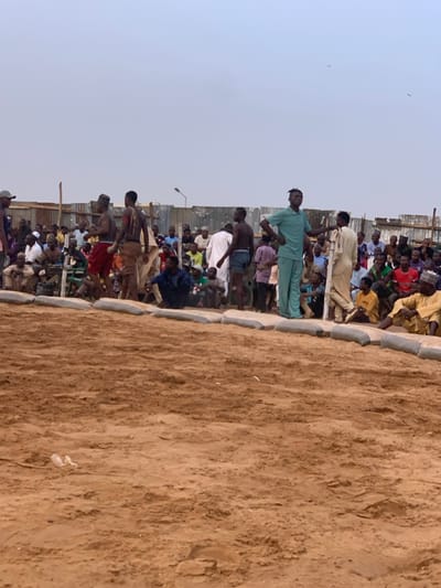 The image shows a group of people gathered in an open, sandy area. The sand is light brown and appears to be dry. There are several people standing around a dirt pit, possibly a wrestling pit, while other people are sitting and watching. A group of men in the center are dressed in traditional clothing and appear to be preparing for a wrestling match. Some of the men have their shirts off and are wearing shorts or loose trousers. Some of the men are holding sticks or other objects that could be used in the match. The scene is located in the city of Sokoto, Nigeria, at latitude 13.049 and longitude 5.238. There are several structures in the background, possibly buildings or fences, made of concrete or cinder blocks. There is a streetlight visible in the background as well. A dusty, grey sky is above the scene. There are several birds flying in the sky. The scene is typical of a wrestling match in Sokoto, Nigeria, where many locals attend and participate in this traditional sport.