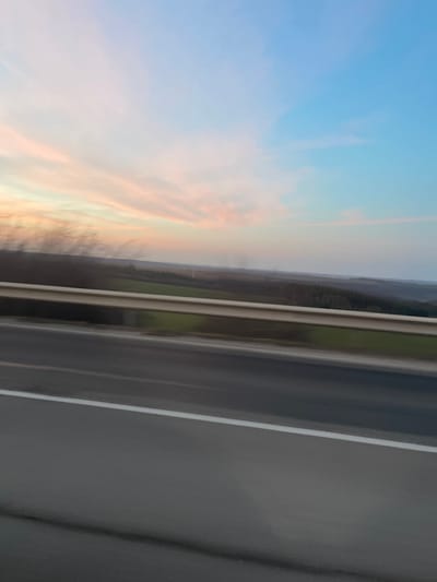 A panoramic landscape image captured from a moving vehicle in the vicinity of Osenets, Bulgaria, likely during dusk or dawn, is presented. The foreground features a grey asphalt road with a solid white lane marking on the right edge, overlaid by a light-colored metal highway guardrail, both exhibiting significant motion blur. Blurred dark vegetation is present on the extreme left. The mid-ground displays an expansive, undulating terrain composed of green agricultural fields and dense, dark forested areas. The background is dominated by a sky gradient, transitioning from soft pink and orange wispy clouds on the left to a clear light blue on the right. Faint, dark plumes or haze are discernible on the distant horizon. No individuals or other specific objects are identifiable.