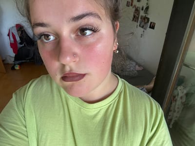 A young woman is posing for a close-up selfie. She is wearing a light green t-shirt and has dramatic makeup on, including false eyelashes and dark lipstick. Her hair is pulled back, and she has a nose stud and earrings. The background is blurry but appears to be a room with photos on the wall and clothing items. The image quality is slightly grainy. There is no information in the image to suggest the location is Lörrach, Germany.