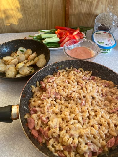 In an indoor setting, likely a kitchen or dining area, a meal is being prepared or served. The scene is laid out on a patterned tablecloth. In the foreground, a large frying pan is filled with macaroni pasta mixed with sliced sausage and covered in a creamy cheese sauce. To the left, another frying pan contains sliced, pan-fried potatoes, some of which are topped with chopped green onions. Behind these pans, a white plate holds a selection of fresh vegetables, including sliced cucumbers, tomatoes, red bell pepper strips, and green onions. To the right of the vegetables, a small glass bowl contains a pinkish sauce, possibly a dip or dressing. Further to the right, a cylindrical plastic container with a blue lid is visible, labeled in Cyrillic with "Svežá" and other text, indicating a food product, likely dairy. Above this container sits a decorative glass jar with a lid, containing dark, small items. The background features a light brown, wooden-paneled wall. The lighting suggests natural light, possibly from a window, casting a warm glow and shadows on the wooden wall. The overall impression is of a home-cooked meal being presented, with ingredients and dishes arranged for consumption. The city of Soroca, Moldova, is not explicitly depicted or indicated by any visual cues in the image.