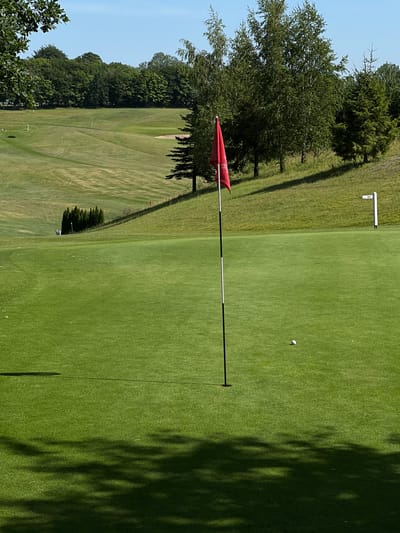 A golf course is pictured on a clear day in Kawle Górne, Poland. The foreground features a vibrant green putting green with a golf ball and a flagstick with a red flag positioned near a hole. In the mid-ground, the rolling hills of the golf course are covered in manicured grass, dotted with trees and a sand trap. Further in the distance, a dense line of trees forms a backdrop against the bright blue sky. The scene is bathed in sunlight, casting shadows across the grassy terrain.
