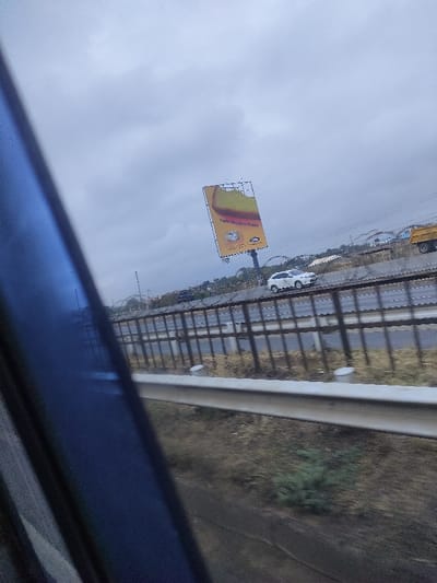 A cloudy view from a moving vehicle, possibly a bus or train. A highway is visible beyond a guardrail. On the highway, a white car and a yellow truck are seen. A large billboard advertising "Taste Hazelnut Roast" stands tall on the side of the road. The roadside vegetation appears dry. A dark, blurry object occupies the left foreground, obstructing some of the view.