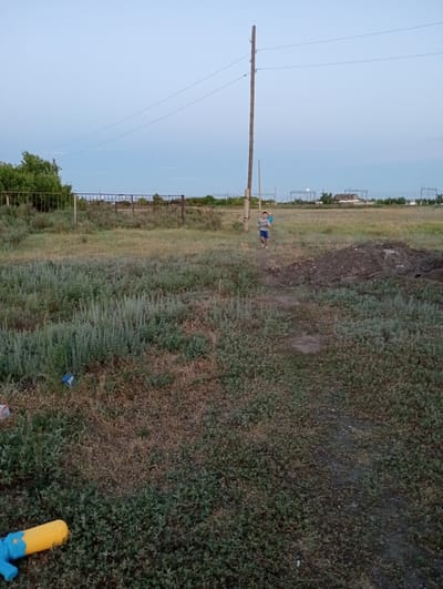 The image depicts an outdoor scene in the city of Октябрьское, Kazakhstan, likely during twilight or early evening, given the soft, diffused light and clear sky. In the foreground, a yellow and blue water gun is visible on the ground amidst green and brown vegetation. A dirt path meanders through a grassy field. Further back, a young boy wearing a grey t-shirt and blue shorts is running towards the right side of the frame. Behind him, another child is visible in the distance. A tall wooden utility pole with power lines runs vertically through the scene, dominating the mid-ground. A metal fence encloses an area with trees and more vegetation on the left side of the image. In the background, structures suggestive of buildings and electrical infrastructure are present, along with more trees and a light-colored, possibly white, object in the sky which might be the moon or a distant building. The terrain appears somewhat uneven with a pile of dark soil to the right of the path.