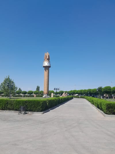 A tall, slender, cream-colored monument stands prominently in the center of the frame under a clear blue sky. The monument has a decorative, silver band around its upper portion, adorned with intricate patterns. Above the band, it tapers to a point, topped with a small cross. To the left of the monument, a mature green tree stands, with a black lamppost beside it. A paved pathway, made of large, square gray stones, leads from the foreground towards the monument. On the left side of the pathway, a black, empty wheelchair is parked near a meticulously trimmed green hedge. This pathway is flanked by the same type of green hedge on both sides, creating a formal garden setting. In the background, a row of neatly shaped green trees forms a visual boundary, with more trees and some indistinct structures visible further beyond. The overall scene depicts a well-maintained outdoor public space, possibly a park or memorial site, in Artashat, Armenia.