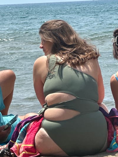 A sunlit outdoor scene shows a woman sitting on a sandy beach by the sea. The woman has brown hair and is wearing a green swimsuit. A multicolored towel is draped next to her. A person in blue shorts sits adjacent, holding a mobile phone. The location is likely a beach in Ortaca, Turkey, based on visual cues like the sea, sand, and climate.