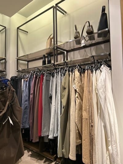 The image captures an interior view of a clothing store, likely a fashion retail outlet in Chalkida, Greece. The scene is dominated by a well-organized display of apparel and accessories.
On the right side, numerous pairs of trousers or pants are neatly arranged on black hangers along a metal clothing rail. The garments exhibit a deliberate color gradient, transitioning from light neutral tones like white and cream at the top, through various shades of beige, khaki, and olive green, and then descending into darker colors including different grays, a prominent red, maroon, blue, and dark brown. This color-coordinated arrangement highlights an intentional retail merchandising strategy.
To the left of the clothing rack, a modern shelving unit constructed with dark metal frames and glass shelves extends vertically. On the top shelf, three handbags are displayed: a black hobo bag and two smaller, structured bags in light brown or khaki. A small white tag is visible on one of these handbags, which is unreadable. Further down, on a lower glass shelf, another beige handbag is presented alongside two pairs of dark-colored boots, each placed on a shoe stand.
The store's interior features light-colored walls, and the overall lighting suggests an indoor environment with artificial illumination. No people are visible, emphasizing the merchandise and the retail display layout. The display elements appear modern and minimalist.