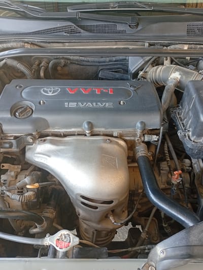 This is a detailed, close-up, eye-level, medium shot of a car engine bay, specifically focusing on the top portion of an engine. The engine appears to be from a Toyota vehicle, indicated by the Toyota logo and "VVT-i 16 VALVE" inscription on a black engine cover. Other visible components include various hoses, wires, a silver heat shield, and part of the air intake system. The engine is dusty and appears to be in a state of disuse or undergoing maintenance, given the lack of activity. The background consists of other engine components and the underside of the car's hood. There are no people present. The context of Seýdi, Turkmenistan, is not visually represented in the image itself; it's an external piece of information.