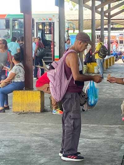 The image, taken in San José de Barlovento, Venezuela, shows a crowded bus station platform. A man wearing a brown baseball cap, maroon tank top, grey pants, and black and red sneakers stands in the foreground, holding a blue plastic bag. He is interacting with someone just out of frame. A group of people are waiting near a green and white bus. Some are boarding. Yellow concrete blocks are scattered on the platform. The ground is grey and uneven. A covered structure provides shade above. Other buses and people are visible in the background, indicating a busy transportation hub.