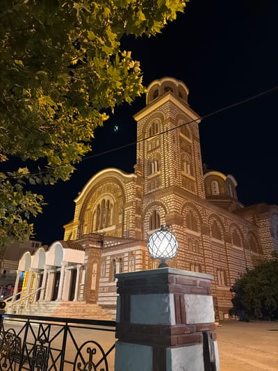 At night in Nea Kallikratia, Greece, a lit church is visible. The church is made of brick and stone, with arched windows and a tall bell tower. In the foreground, a decorative lamp sits on a brick and stone pedestal. A black metal fence runs along the bottom of the image. Trees with green leaves are visible on the left and right sides of the church. The sky is dark.