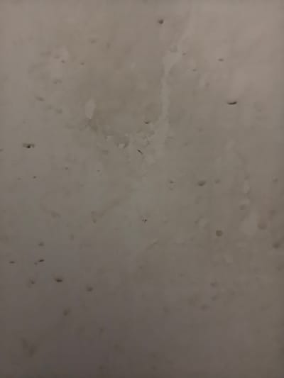 The image is a close-up, textured view of a light-colored, possibly concrete or plaster surface. It features numerous small, dark specks and irregular, lighter patches, suggesting dirt, water stains, or imperfections in the material. The lighting is dim, creating subtle shadows and variations in tone. There are no discernible people, objects, or actions depicted. The scene appears to be a wall or ceiling surface. The provided location of Ново-Талицы, Russia, does not offer specific visual context that can be confirmed from the image itself.