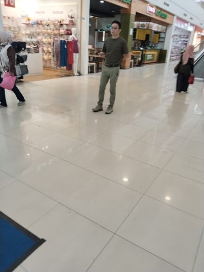 The image shows a man wearing a dark t-shirt and olive green pants standing in a well-lit shopping mall. He is wearing glasses and has short dark hair. In the background, to the left, a woman wearing a light-colored hijab and a dark jacket is walking away, carrying a pink bag. To the right, another woman in a pink hijab and dark clothing is walking away, also carrying a bag. Various shop displays are visible, including one with lingerie and mannequins. The floor is tiled with large, light-colored tiles, reflecting the overhead lighting. The overall environment suggests a public commercial space, likely a mall in Kudus, Indonesia, given the presence of a store with what appears to be Indonesian signage.