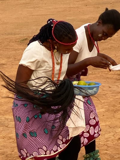 This media file captures two women seated, dressed in traditional attire, engaged in what appears to be a cultural or ceremonial activity in Agunu, Nigeria. The image is oriented vertically but depicts the women in a seated posture.
The woman in the foreground, positioned to the lower left, has dark braided hair styled with a red beaded headband. She wears a white short-sleeved top and a long, multi-strand red beaded necklace. A distinct gold-colored earring with a circular disc and a smaller pendant adorns her right ear. Her lower body is covered by a vibrant purple fabric patterned with stripes and stylized teal shell-like motifs. Long, dark, straight hair, possibly extensions or a wig, cascades over her lap and the patterned fabric.
Behind her, slightly to the upper right, sits another woman. Her dark hair is pulled back, and she also wears a white short-sleeved top and a red beaded necklace. Notable white markings, possibly chalk or paint, are visible on her cheek. She holds a piece of white cloth or paper in her hands, appearing to examine or manipulate it with focus. Her lower body is covered by a maroon fabric featuring white and purple shell-like patterns.
Between the two women, on the ground, rests a small blue plastic basket containing several green, spherical fruits, likely limes or small oranges. The setting appears to be indoors or under a shaded structure, as indicated by the light brown, possibly wooden or earthen floor. The lighting suggests it is daytime. The women's expressions are neutral and focused, hinting at the solemnity or importance of their activity. There is no visible text in the image.