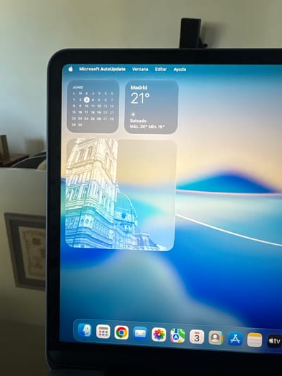 A digital device screen, possibly a tablet or laptop, is shown in a portrait orientation, displaying its desktop interface. The screen's background is an abstract wallpaper with soft blue, yellow, and white hues.
On the left side of the screen, a menu bar is visible at the top, showing "Microsoft AutoUpdate" followed by standard menu options in Spanish: "Ventana" (Window), "Editar" (Edit), and "Ayuda" (Help), alongside an Apple logo. Below this, two widgets are displayed. The left widget shows a calendar for "JUNIO" (June), with the 3rd highlighted, indicating the current date. The right widget is a weather display for "Madrid", showing "21°" as the current temperature under "Soleado" (Sunny) conditions, with a maximum of "30°" and minimum of "16°". Next to these, another widget displays a stylized image of a classical architectural building, possibly the Duomo in Florence.
Along the right edge of the screen, a dock of application icons is present. Visible icons include Apple TV (with "tv" text), Notes, App Store, Camera, Calendar (showing "Mié 3" for Wednesday 3rd), Maps, Photos, Mail, Google Chrome, an App Library icon, and another application icon partially visible at the bottom.
The device itself has a black bezel. In the physical background, a light-colored wall is seen, with a dark, indistinct object protruding from the left side, possibly a stand or cable. Below the screen, a framed picture or object is partially visible. The Spanish text throughout the interface confirms a Spanish-speaking user or region, consistent with the provided location context of Spain. The widgets suggest a warm, sunny day in Madrid, with the date being June 3rd.