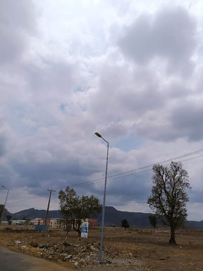 The image depicts an expansive, dry landscape in Rafin Pa, Nigeria, under a heavily clouded sky with a small visible patch of blue. In the foreground, an unpaved dirt road is visible on the left, transitioning to reddish-brown, uncultivated ground with scattered rocks and debris. Two streetlights are present: a prominent grey metallic pole with a luminaire is slightly right of center, and another shorter pole with a luminaire is on the far left. At least two wooden utility poles with multiple black lines span the mid-ground. A white sign with "25 KPH" and "SPEED LIMIT" is visible on a pole near the center-left, alongside a vertical banner-like sign. Vegetation includes a large, mature tree on the right and a smaller, bushy tree left of center. In the mid-ground, multiple light-colored, multi-story buildings are situated at the base of distant hills or mountains that form the horizon. No individuals are visible, and no active human interactions are depicted.