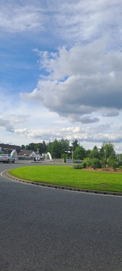 The image shows a circular grassy roundabout in the foreground, with an asphalt road curving away from it to the left. In the middle ground, there are two white arch bridges crossing over what appears to be a river or canal. Beyond the bridges, there are trees and some buildings, including a multi-story building with blue accents on the left. A silver car is visible on the road to the left of the roundabout. The sky above is partly cloudy with blue patches. The scene is likely in Castlebar, Ireland, given the presence of the arch bridges which are a notable feature of the town.