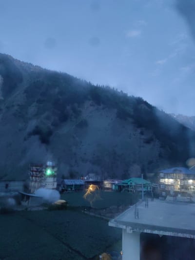 This is a photograph of a mountainous landscape with a village nestled at the foot of a slope. The image appears to have been taken at dusk or during a period of low light, as indicated by the muted colors and the artificial lights emanating from some buildings. The sky is overcast, and there is a noticeable mist or fog obscuring the upper slopes of the mountain, which is covered in sparse vegetation and trees.

In the foreground, there is a dark, cultivated field, possibly farmland. Several buildings are visible in the midground. One prominent structure is a multi-story building with illuminated windows, suggesting it is a hotel or residential building. Next to it, there's a building with a green roof. To the left, another tall building with several floors and lit windows is visible. Smaller structures and what appears to be a greenhouse or a covered farm area are also present.

The overall scene depicts a rural or semi-rural settlement in a mountainous region. The lack of distinct human figures or specific cultural landmarks makes it difficult to pinpoint a precise location without additional context. This image is not of Udaipur, India, as Udaipur is located in a desert region and is known for its lakes and palaces, not mountainous terrain and mist.