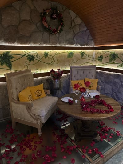An intimate dining setup includes a round wooden table adorned with rose petals forming a heart shape. Two luxurious chairs with yellow logo pillows face each other. Red rose petals are scattered around the table and across a transparent glass floor revealing the space below. A floral arrangement sits between the chairs. A stone wall with a decorative wreath and grape vine pattern adds to the romantic ambiance.