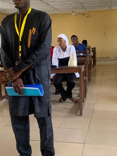 This image depicts an indoor academic or lecture setting within an educational institution in Abobo, Nigeria. In the immediate foreground, a person, likely male, stands with their arms crossed at the waist, holding a two-toned blue folder or notebook. They wear a dark garment patterned with small white polka dots and a yellow lanyard around their neck, which displays "POLYTECHNIC" text and holds pens in a brown-accented pocket. In the midground, a female is seated at a wooden desk, wearing a white head covering, a white long-sleeved shirt over a black top, and black trousers. Her legs are crossed at the ankles, and she wears patterned sandals. On her desk sits a light-colored, possibly cream, structured handbag and a dark object, likely a smartphone. Her gaze is directed towards the left side of the frame. Behind her, other students are visible seated at identical wooden desks, including a male in a blue top in the adjacent row looking forward. The room features light yellow walls, a tiled floor, and a white ceiling equipped with a white ceiling fan and a small black CCTV camera.