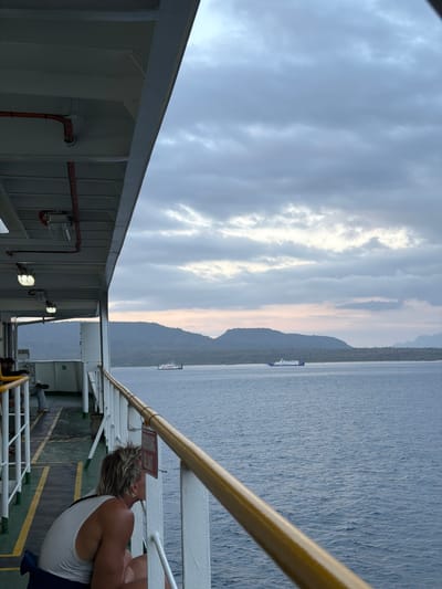 This is a medium shot of a person on the deck of a ferry, looking out at the sea. The person is wearing a white tank top and has blonde hair. They are sitting on the deck and leaning against the railing. In the background, there is a body of water and a distant island. A ferry boat is visible in the distance. The sky is cloudy and overcast. The location is Melaya, Indonesia.