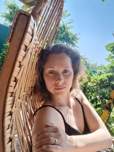 A woman is pictured in a close-up shot, taken outdoors during the day. She is seated in a woven chair, leaning against its back. The woman has light brown, wavy hair and is looking directly at the camera. She is wearing a black top and a ring on her left hand, which is positioned across her chest. The background is filled with lush green foliage and a clear blue sky. The scene appears to be set in a garden or natural environment, possibly within the city of Maslova Pristan', Russia, suggested by the presence of greenery and a relaxed outdoor setting typical of residential areas.