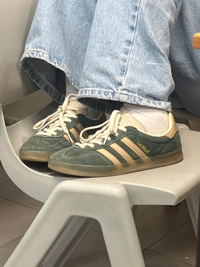 This image depicts a close-up view of a person's lower legs and feet, resting on a light grey plastic chair in an indoor setting within Ness Ziona, Israel. The individual wears light blue denim jeans, white ankle socks, and a pair of dark green suede Adidas Gazelle sneakers with light tan stripes and heel tabs. The shoes feature off-white laces and translucent, amber-toned rubber soles. "GAZELLE" is embossed in gold lettering on the lateral side of the right shoe. The floor beneath the chair appears to be tiled. No other individuals or specific environmental details beyond the chair and floor are clearly discernible.