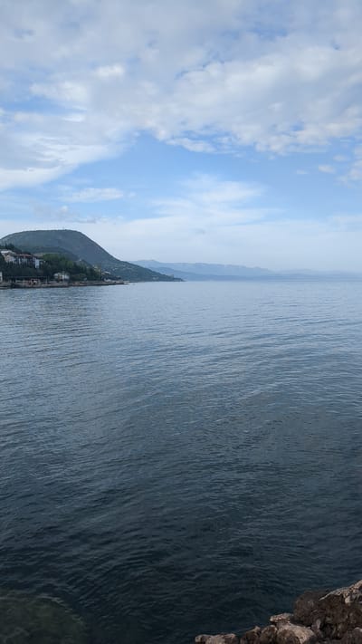 The image shows a body of water, possibly the Black Sea, with a coastline visible in the lower left. The coastline features a hillside with some vegetation and a few buildings clustered along the shore. The sky is blue with scattered white clouds. The right side of the image includes a rocky outcropping, possibly a cliff. The location is identified as Bondarenkovo, Ukraine, but there are no people or specific actions visible in the image. The primary subjects are the water, the coastline, and the sky.