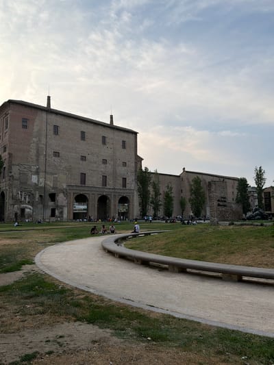 The image shows the exterior of a large, old brick building with arched entrances on the ground floor, located in Parma, Italy. The building appears somewhat weathered and shows signs of age. In front of the building, there is a grassy area where several people are sitting and lying down, enjoying the outdoors. A curved, sandy pathway winds through the grass, bordered by a concrete bench. Tall, slender trees are planted in a line behind the pathway and in front of a portion of the building. In the background, another section of the building stretches horizontally, and further beyond, there are more trees and a glimpse of other structures. The sky is partly cloudy, with a pale blue hue. The overall scene depicts a public park setting with historical architecture.