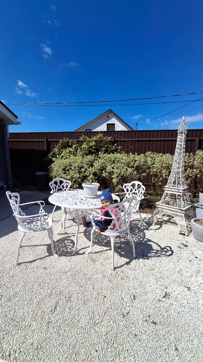 The image depicts an outdoor setting in what appears to be a backyard or patio area. A child is seated at a white metal table with ornate chairs. Behind the table and chairs, there's a brown fence or wall and a patch of greenery, possibly bushes or shrubs. A white Eiffel Tower replica stands behind the greenery. The ground is covered with small stones or gravel. The sky is blue with some clouds. The location might be Oliki, Russia, based on the question.
