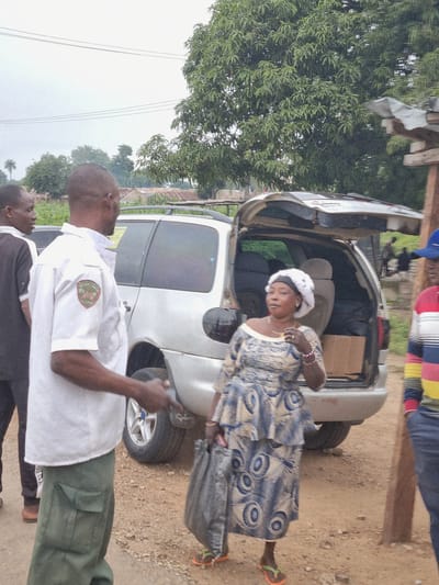 In the city of Marke, Nigeria, a group of people are gathered around a silver minivan with its rear hatch open. A man in a white short-sleeved uniform shirt with a patch on the left breast and green cargo pants stands in the foreground, facing away from the camera and gesturing with his left hand. To his left, another man in a black shirt and dark pants is partially visible. A woman in a blue and white patterned dress and a white head covering is standing to the right of the minivan, holding a dark bag. Her right hand is near her face, and she is wearing a necklace and bracelets. Behind her, a person in a striped shirt and blue pants is visible. The ground is covered in gravel and dirt, and vegetation and buildings are visible in the background under an overcast sky.